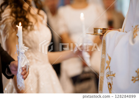 Orthodox Christian Wedding Ceremony: Lighting Candles, Priest Vestments, White Dress, Religious Ritual, Gold Embroidery, Soft Focus, Indoor Scene. 128284899