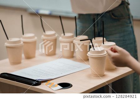 Beverage Sampling Event: Array of Paper Cups and Hand Reaching, Light Wood Table, Denim Jeans, Blank Paper, Neutral Palette, Testing Scenario. 128284921
