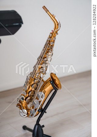Golden Alto Saxophone on Stand, Music Instrument, Studio Shot, Warm Lighting, Brass Texture, Jazz Performance, Solo Practice, Elegant Design. Golden Alto Saxophone on Stand, Music Instrument, Studio Shot, Warm Lighting, Brass Texture, Jazz Performance, Solo Practice, Elegant Design. 128284922