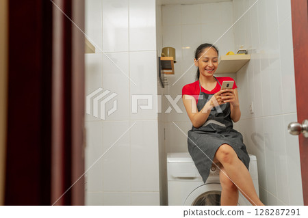 Young Woman Relaxing on Washer, Using Smartphone in Modern Bathroom Setup Young Woman Relaxing on Washer, Using Smartphone in Modern Bathroom Setup 128287291