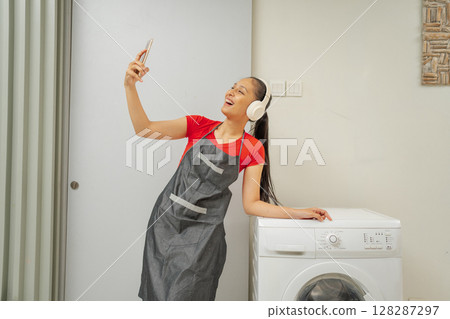 Cheerful Young Woman Happily Taking a Selfie in a Bright Modern Laundry Room Cheerful Young Woman Happily Taking a Selfie in a Bright Modern Laundry Room 128287297