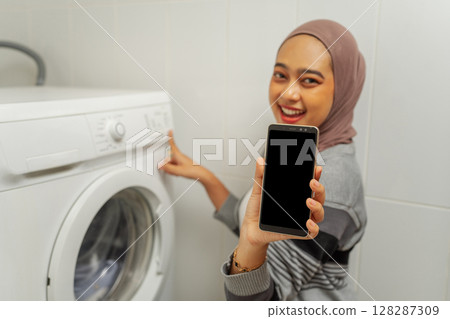 A Happy User is Showcasing Their Smartphone Alongside a Washing Machine at Home 128287309