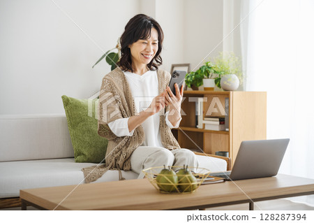 A middle-aged woman in her 50s looking at her smartphone in the living room at home A middle-aged woman in her 50s looking at her smartphone in the living room at home 128287394