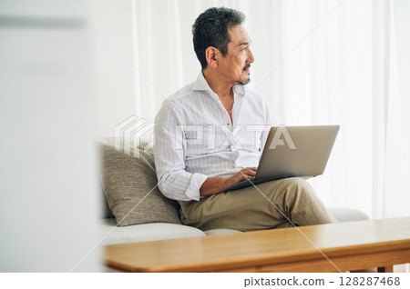 A middle-aged man in his 50s working on a computer on the sofa in the living room A middle-aged man in his 50s working on a computer on the sofa in the living room 128287468