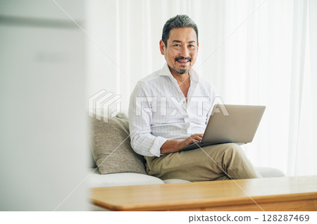 A middle-aged man in his 50s working on a computer on the sofa in the living room A middle-aged man in his 50s working on a computer on the sofa in the living room 128287469