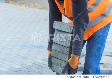 Laborer in an orange vest lifts several brick blocks to install paving stones in residential walkway. Laborer in an orange vest lifts several brick blocks to install paving stones in residential walkway. 128290535