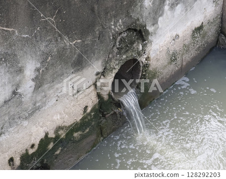Small pipe embedded in weathered concrete wall releases water into murky water body, with moss and algae growth around pipe opening, creating natural, aged, and slightly neglected scene Small pipe embedded in weathered concrete wall releases water into murky water body, with moss and algae growth around pipe opening, creating natural, aged, and slightly neglected scene 128292203