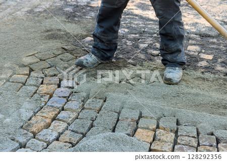 Construction worker carefully positions cobblestones amidst fresh gravel on busy city street. Construction worker carefully positions cobblestones amidst fresh gravel on busy city street. 128292356