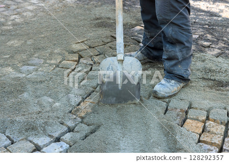 Construction worker uses shovel to spread gravel over cobblestones at building site 128292357
