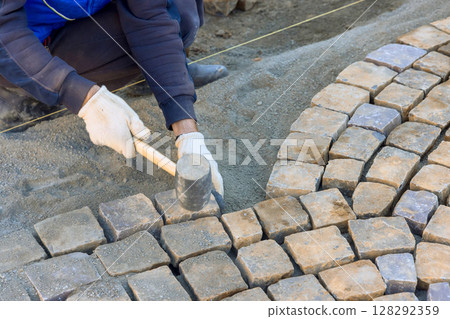 Worker meticulously arranging cobblestones on pathway, demonstrating skilled craftsmanship under works day Worker meticulously arranging cobblestones on pathway, demonstrating skilled craftsmanship under works day 128292359