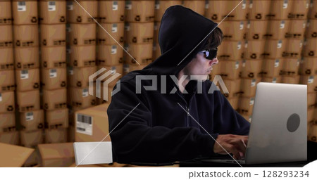Image of hacker using laptop with stacks of boxes in warehouse 128293234