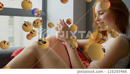 Young woman using smartphone at home surrounded by floating emojis, looking amused 128293466