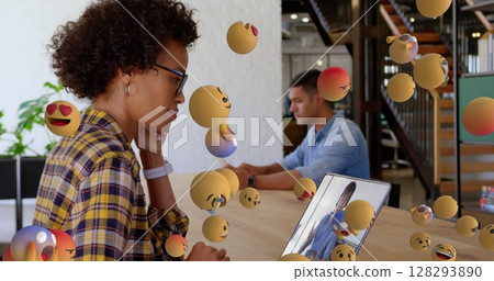 African American woman using laptop with floating emojis in modern office 128293890