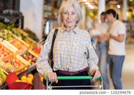 In food store, senior woman with trolley looking for something on shelf of showcase In food store, senior woman with trolley looking for something on shelf of showcase 128294676