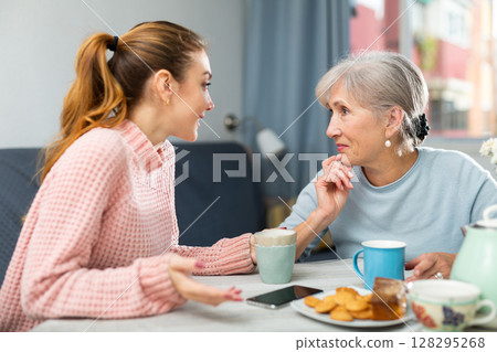 Mother and granddaughter talking in kitchen and drinking coffee 128295268