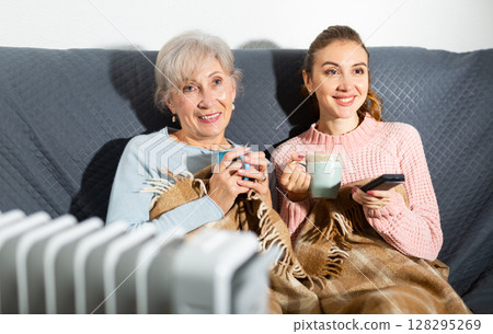 Happy elderly woman with adult daughter drinking tea and watching movie Happy elderly woman with adult daughter drinking tea and watching movie 128295269