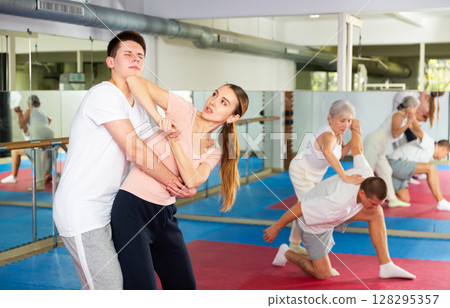 Young woman trying to break free of back grip of male opponent, elbowing to chin Young woman trying to break free of back grip of male opponent, elbowing to chin 128295357
