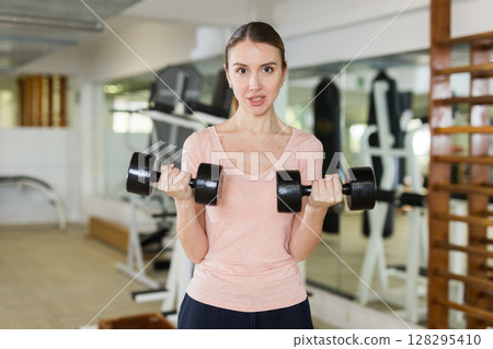Portrait of positive woman with dumbbells in his hands in gym 128295410