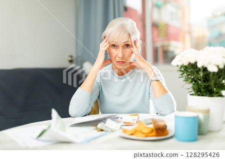 Upset senior woman looking worriedly at papers at home table 128295426