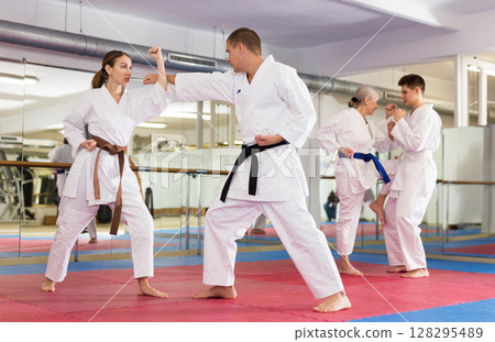 Man and woman in kimono and belt fighting with opponent during group karate training in gym Man and woman in kimono and belt fighting with opponent during group karate training in gym 128295489