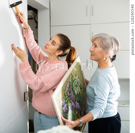 Young woman helping elderly mother to hang painting on wall 128295540