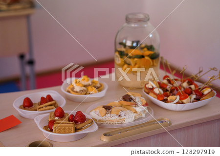 Festive Food Spread with Jar of Pickles, Fruit Skewers, Desserts and Savory Bites on Pink Table, Bright Interior Scene Festive Food Spread with Jar of Pickles, Fruit Skewers, Desserts and Savory Bites on Pink Table, Bright Interior Scene 128297919