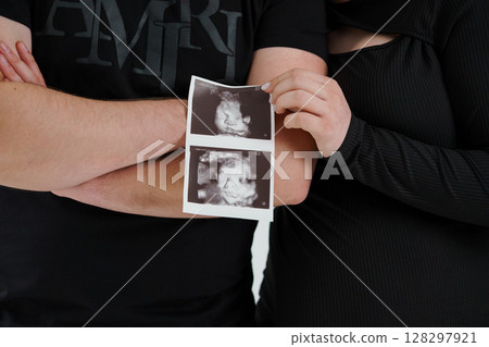 Couple Holding Ultrasound Image of Baby. Black Clothing. Excited Expectant Parents. Pregnancy Announcement. Close-Up Shot. Dark Background. Emotional Moment Couple Holding Ultrasound Image of Baby. Black Clothing. Excited Expectant Parents. Pregnancy Announcement. Close-Up Shot. Dark Background. Emotional Moment 128297921