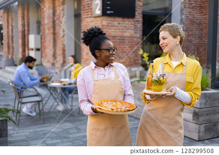 Two waitresses smiling, carrying pizza and drinks outdoors at a restaurant, with customers in the background. Two waitresses smiling, carrying pizza and drinks outdoors at a restaurant, with customers in the background. 128299508