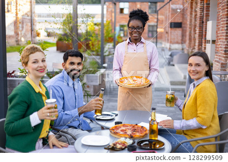 A smiling waitress serves pizza to a group of friends at an outdoor cafe. A smiling waitress serves pizza to a group of friends at an outdoor cafe. 128299509