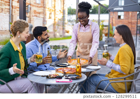 A cheerful server presents pizza to a group of friends enjoying drinks at an outdoor restaurant. A cheerful server presents pizza to a group of friends enjoying drinks at an outdoor restaurant. 128299511