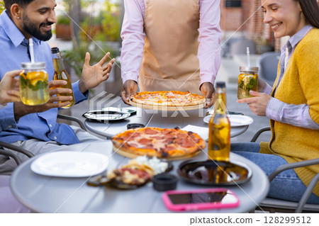 A diverse group of people enjoying pizza, drinks, and each other's company outdoors. A waiter is serving pizza. A diverse group of people enjoying pizza, drinks, and each other's company outdoors. A waiter is serving pizza. 128299512