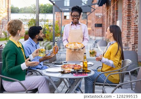A smiling waitress serves pizza to a group of friends at an outdoor cafe, enjoying drinks and the sunny day. 128299515