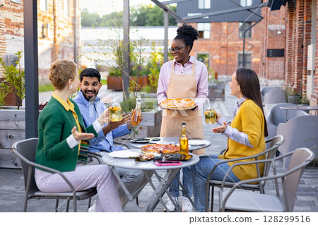 A waitress serves pizza and drinks to a group of friends at an outdoor restaurant. They are enjoying their time. 128299516