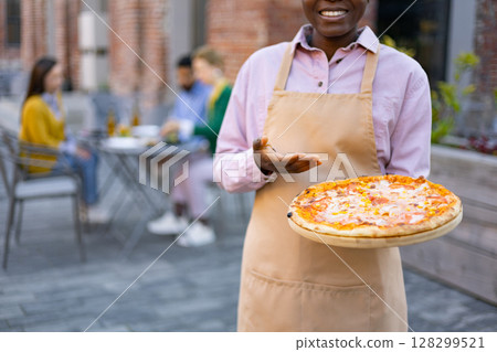 A smiling waiter presents a delicious pizza on a wooden board, ready to be served to customers enjoying an outdoor dining experience. 128299521