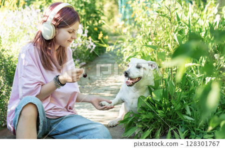 Teenage girl wearing headphones sharing cherries with her dog in garden 128301767