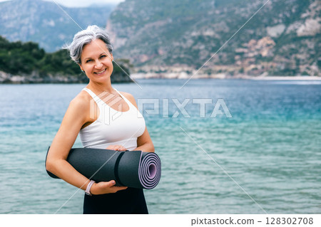 Happy senior woman holding yoga mat by the sea, symbol of active lifestyle and graceful aging 128302708