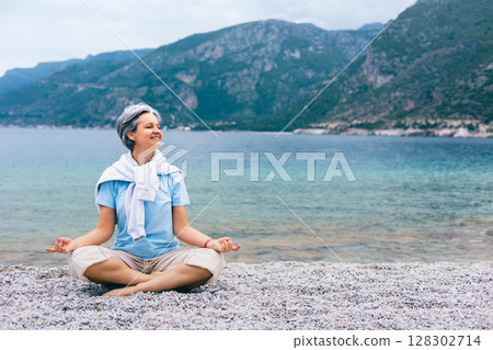 Happy mature woman meditating by the sea, enjoying peaceful moment on rocky beach outdoors 128302714