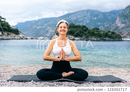 Senior woman in Easy Pose doing Namaste gesture on beach with ocean and mountain background 128302717