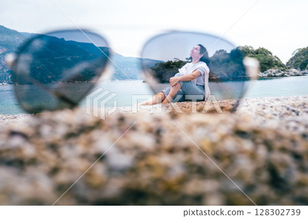 Dreamy beach view of smiling woman through sunglasses, relaxing near sea and forested cliffs 128302739