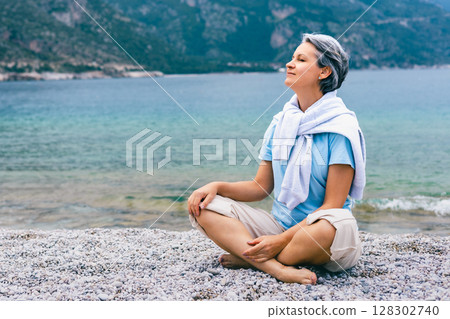 Mature woman meditating near ocean, enjoying tranquility and fresh air on mountain beach 128302740
