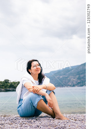 Young woman sitting on rocky beach and enjoying peaceful coastal view on a cloudy summer day 128302749