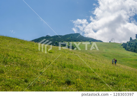 Sunny summer meadow hike on green mountain slope under bright blue sky and clouds. High quality photo 128303397