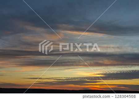Colorful sunset over the horizon with clouds dispersing in various shades of orange and blue near a tranquil landscape Colorful sunset over the horizon with clouds dispersing in various shades of orange and blue near a tranquil landscape 128304508