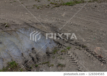 Tire tracks in muddy ground near a small puddle reflecting surrounding vegetation during a sunny day in early spring 128304565