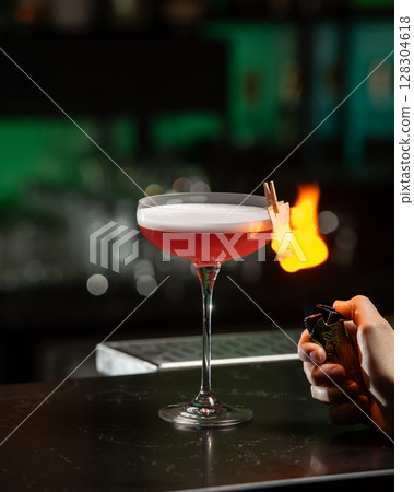 A dramatically lit, crimson cocktail with a white foam top is being lit by a bartender, creating a burst of flame from a citrus peel garnish clipped to the glass 128304618