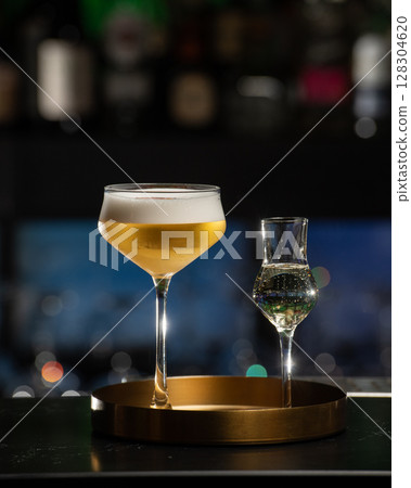 A beautifully presented golden-hued cocktail with a rich foam top is served alongside a small shot glass of clear liquid, all resting on a chic golden tray 128304620