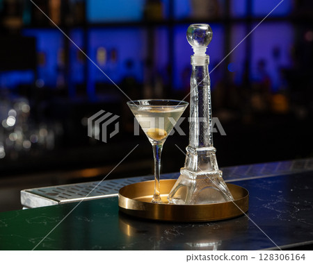 A martini glass with an olive and a clear stylish shaped decanter, both containing an alcoholic cocktail, are presented on a golden tray on a bar counter with a blurred blue background A martini glass with an olive and a clear stylish shaped decanter, both containing an alcoholic cocktail, are presented on a golden tray on a bar counter with a blurred blue background 128306164