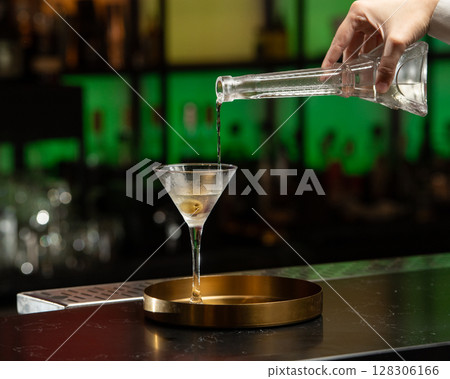 A clear alcoholic cocktail, garnished with an olive, is being poured into a martini glass from a stylish decanter, all set on a golden tray at a bar A clear alcoholic cocktail, garnished with an olive, is being poured into a martini glass from a stylish decanter, all set on a golden tray at a bar 128306166