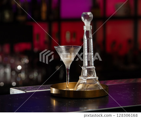 A classic martini in a frosted glass stands beside a unique Eiffel Tower-shaped decanter, both presented on a golden tray 128306168