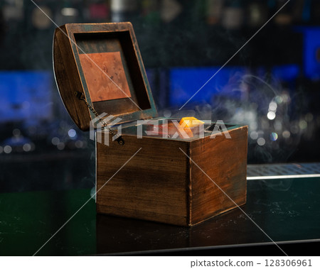 A smoky, dark red alcoholic cocktail with ice and an orange peel garnish is presented in a rocks glass nestled inside an open wooden box. 128306961